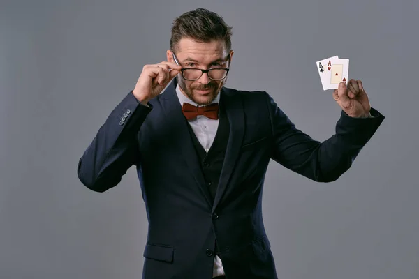 Man in black classic suit, red bow-tie, glases is showing two playing cards, posing on gray studio background. Gambling, poker, casino. Close-up. — ストック写真