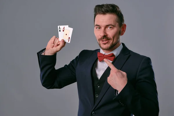 Man in black classic suit and red bow-tie showing two playing cards while posing against gray studio background. Gambling, poker, casino. Close-up. — Stockfoto