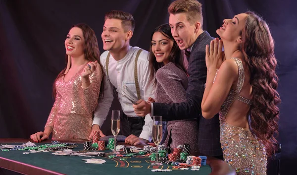 Friends playing poker at casino, at table with stacks of chips, money, cards, champagne on it. Celebrating win, excited. Black background. Close-up. — Φωτογραφία Αρχείου