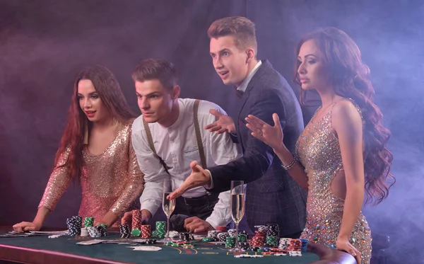 Friends playing poker at casino, at table with stacks of chips, money, cards, champagne on it. Celebrating win, excited. Black background. Close-up. — Stock fotografie