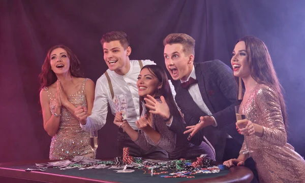 Friends playing poker at casino, at table with stacks of chips, money, cards, champagne on it. Celebrating win, excited. Black background. Close-up. — Φωτογραφία Αρχείου