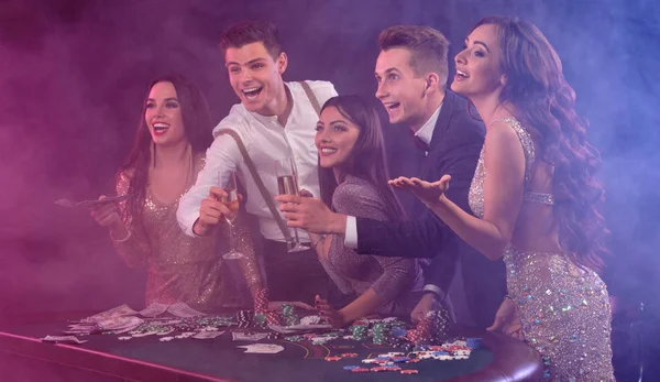 Friends playing poker at casino, at table with stacks of chips, money, cards, champagne on it. Celebrating win, excited. Black background. Close-up. — ストック写真