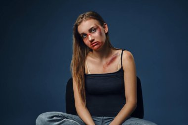 Blonde girl with bruises on her face and chest posing sitting on blue studio background. Domestic violence, abuse. Close-up, copy space.