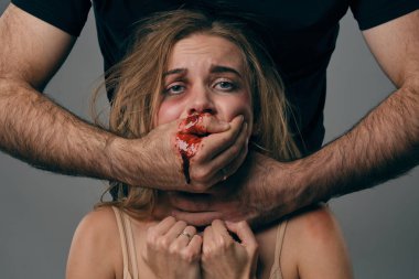 Strong man strangling scared female with bruises on her face. Blood from her mouth flows down his arm. Gray background. Domestic violence. Close-up.
