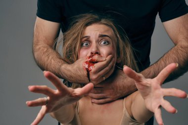 Strong man strangling scared female with bruises on her face. Blood from her mouth flows down his arm. Gray background. Domestic violence. Close-up.
