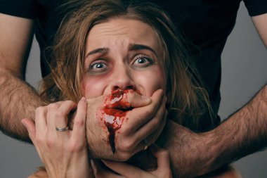 Strong man strangling scared female with bruises on her face. Blood from her mouth flows down his arm. Gray background. Domestic violence. Close-up.