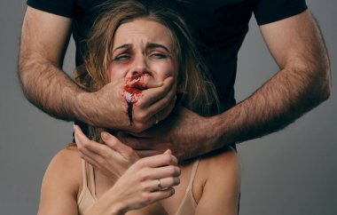 Strong man strangling scared female with bruises on her face. Blood from her mouth flows down his arm. Gray background. Domestic violence. Close-up.