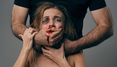 Strong man strangling scared female with bruises on her face. Blood from her mouth flows down his arm. Gray background. Domestic violence. Close-up.