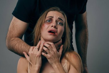 Güçlü adam, yüzünde morluklar olan korkmuş sarışın kadını boğuyor. Gri stüdyo arka planında poz vermek. Aile içi şiddet, taciz. Yakın plan..