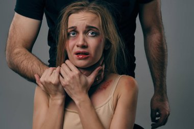 Güçlü adam, yüzünde morluklar olan korkmuş sarışın kadını boğuyor. Gri stüdyo arka planında poz vermek. Aile içi şiddet, taciz. Yakın plan..