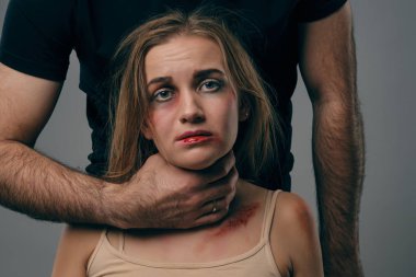 Güçlü adam, yüzünde morluklar olan korkmuş sarışın kadını boğuyor. Gri stüdyo arka planında poz vermek. Aile içi şiddet, taciz. Yakın plan..