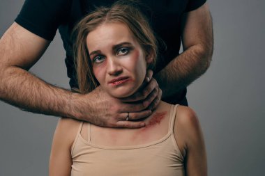 Güçlü adam, yüzünde morluklar olan korkmuş sarışın kadını boğuyor. Gri stüdyo arka planında poz vermek. Aile içi şiddet, taciz. Yakın plan..