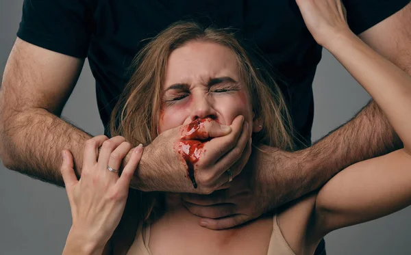 Strong man strangling scared female with bruises on her face. Blood from her mouth flows down his arm. Gray background. Domestic violence. Close-up. — ストック写真