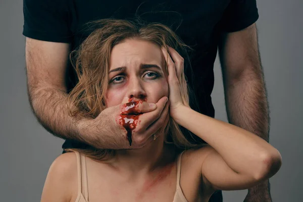 Strong man covered mouth of scared female with bruises on her face. Blood from it flows down his arm. Gray background. Domestic violence. Close-up.