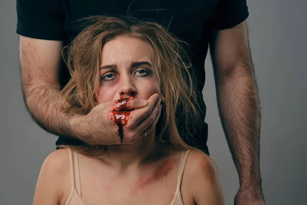 強い男が顔にあざで怖がっている女性の口を覆った。血が彼の腕を流れる。灰色の背景。家庭内暴力。閉鎖. — ストック写真