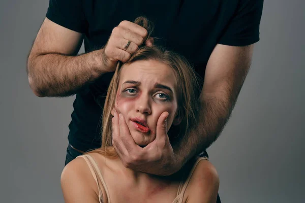 Un hombre fuerte sostiene por el pelo a una mujer asustada con el pelo alborotado y moretones en la cara. Fondo de estudio gris. Violencia doméstica, abuso. Primer plano . — Foto de Stock