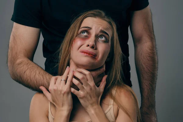 stock image Strong man is strangling scared blonde female with bruises on her face. Posing on gray studio background. Domestic violence, abuse. Close-up.