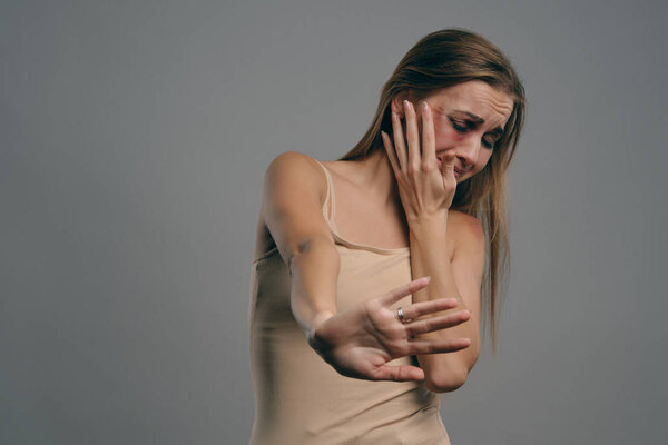 Blonde girl with bruises on her face, trying to protect herself, posing on gray studio background. Domestic violence, abuse. Close-up, copy space.