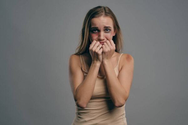 Blonde girl with bruises hiding her face with fists posing against gray studio background. Domestic violence, abuse. Close-up, copy space.
