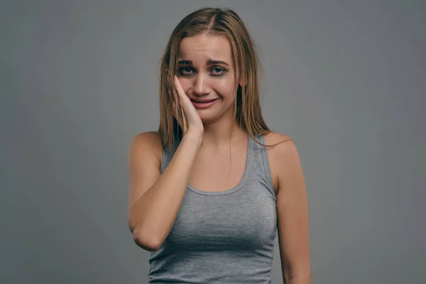Blond meisje met blauwe plekken raakt haar gezicht, haar grijze studio achtergrond. Huiselijk geweld, misbruik. Depressie, wanhoop. Close-up, kopieerruimte. — Stockfoto
