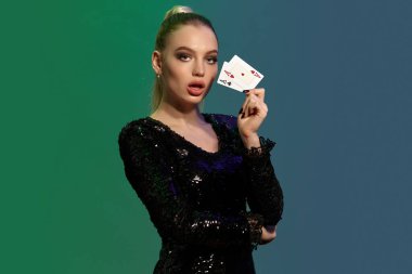 Atkuyruklu, mücevherli ve siyah pullu elbisesi olan güzel sarışın kadın. Ağzını açtı, iki as gösterdi, dirseğini eliyle destekledi, renkli arka planda poz verdi. Poker, kumarhane. Yakın plan.