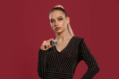 Güzel sarışın kız, at kuyruklu, mücevher takan ve elmaslı siyah elbiseli. Kırmızı arka planda poz veren renkli çipler gösteriyor. Kumar eğlencesi, poker, kumarhane. Yakın plan.