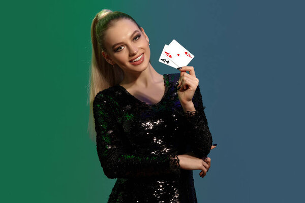 Очаровательная блондинка с хвостиком, в ювелирном и черном платье с блестками. Она улыбается, показывая две игральные карты, поддерживая локоть рукой, позируя на красочном фоне. Покер, казино. Крупный план
