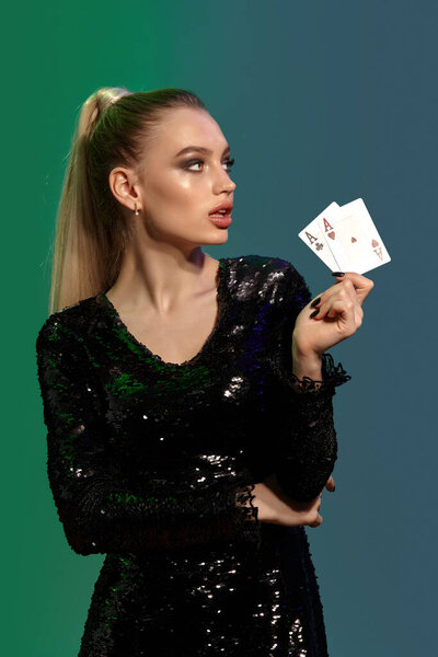 Великолепная блондинка-модель с пони-хвостом, в драгоценностях и черном платье. Она показывает две игральные карты, глядя в сторону, позируя на красочном фоне. Азартные игры, покер, казино. Крупный план
