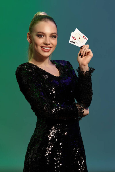 Шикарная блондинка с хвостиком, в ювелирном и черном платье с блестками. Она улыбается, показывая две игральные карты, поддерживая локоть рукой, позируя на красочном фоне. Покер, казино. Крупный план
