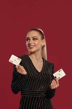 Atkuyruklu, mücevherli ve siyah elmaslı çekici sarışın manken. Gülümsüyor, kırmızı zemine karşı poz verirken iki iskambil kağıdı gösteriyor. Kumar, poker, kumarhane. Yakın plan.