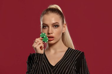 Atkuyruklu, mücevherli ve siyah elmaslı güzel sarışın bayan. Kırmızı arka planda poz veren iki yeşil çip gösteriyor. Kumar eğlencesi, poker, kumarhane. Yakın plan.
