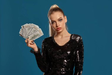 Sarışın, at kuyruklu, mücevherli ve siyah pullu elbiseli bir kadın. Mavi arka planda poz veren yüz dolarlık banknotların hayranı. Kumar eğlencesi, poker, kumarhane. Yakın plan.