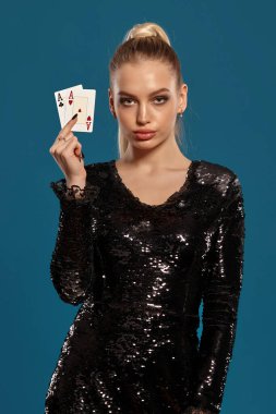 Atkuyruklu, mücevherli ve siyah pullu elbisesi olan güzel sarışın kadın. Mavi stüdyo arka planında poz verirken iki as gösteriyor. Kumar eğlencesi, poker, kumarhane. Yakın plan.