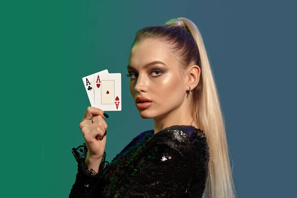 Блондинка-модель с пони-хвостом, в драгоценностях и черном платье. Она показывает два туза, позируя боком на красочном фоне. Азартные игры, покер, казино. Крупный план
