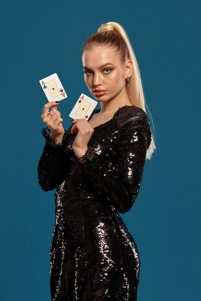 Великолепная блондинка-модель с пони-хвостом, в драгоценностях и черном платье. Она показывает две игральные карты, позируя боком на синем фоне. Азартные игры, покер, казино. Крупный план
