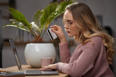 Pembe hırkalı ve gözlüklü sarışın manken. Dizüstü bilgisayarı üzerinde çalışıyor, elinde beyaz plastik kart var. Masada pembe bardakla otururken, avuç içi çömlek içinde. Kapat.