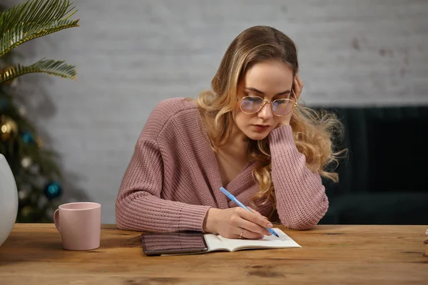 Model blogger in pink cardigan and glasses. Sitting at table, writing in notebook. Palm tree in pot, cup and tablet nearby. Christmas tree. Close up