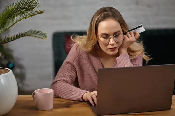 穿着粉色羊毛衫戴眼镜的女士在笔记本电脑上工作，并展示白色塑料卡片。坐在木制桌旁，上有粉红的杯子，手掌插在锅子里。靠近点 — 图库照片