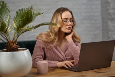 Pembe hırkalı ve gözlüklü sarışın manken. Dizüstü bilgisayarı üzerinde çalışıyor. Pembe bardakla ahşap masada oturuyordum. Avuç içi saksıdaydı. Öğrenci, blogcu. Kapat.