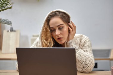 Güzel sarışın kız, blogcu ya da öğrenci, bej kapüşonlu kapüşonlu. Mutfakta, ahşap masada oturup dizüstü bilgisayarına bakıyor. İç mekan. Kapat.