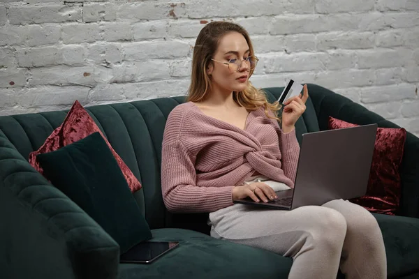 Szőke nő rózsaszín kardigánban és szemüvegben. Zöld kanapén ül, színes párnákkal, műanyag kártyával a kezében, és a laptopját nézi. Közelről. — Stock Fotó