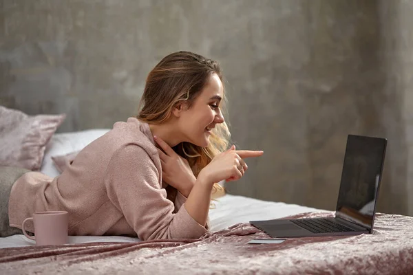 Szőke nő blogger alkalmi ruhában. Mosolyog és a laptopjára mutat, oldalt fekszik az ágyon fehér műanyag kártyával és pohárral. Közelről. — Stock Fotó