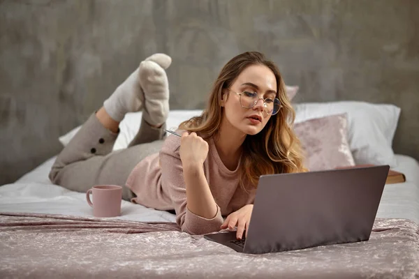 Blogger mulher loira em óculos, roupas casuais. Segurando cartão de plástico, deitado de pernas cruzadas na cama com laptop, livro e copo. Trabalho, educação. Close-up — Fotografia de Stock