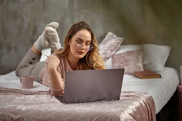 Szőke modell blogger szemüvegben, alkalmi ruhában. Fehér műanyag kártya, laptoppal, könyvvel és csészével az ágyon. Munka vagy oktatás. Közelkép — Stock Fotó