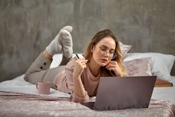 Blogger loira em óculos, roupas casuais. Segurando cartão de plástico branco, deitado na cama com laptop, livro e copo. Close-up, espaço de cópia — Fotografia de Stock