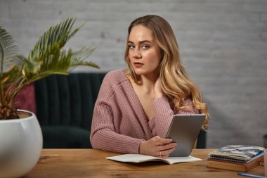 Pembe hırkalı kadın blogcu elinde bir tablet tutuyor. Üzerinde defterler ve saksıda palmiye ağaçları olan ahşap bir masada oturmak. Öğrenci, blogcu. Yakın plan.