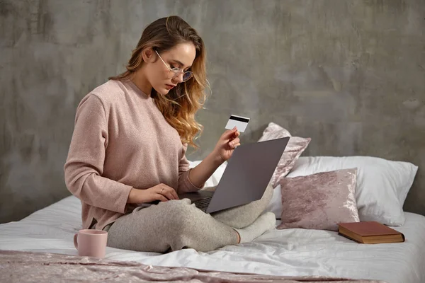 Blogger loira em óculos, roupas casuais. Segurando cartão de plástico e digitando em seu laptop, sentado de lado na cama com livro e xícara. Fechar — Fotografia de Stock