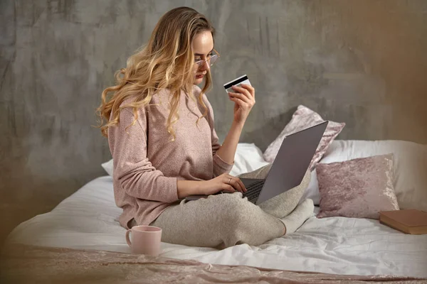 Blogger menina loira em óculos, roupas casuais. Segurando cartão de plástico e laptop em suas pernas, sentado de lado na cama com livro e xícara. Fechar — Fotografia de Stock
