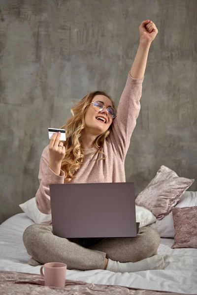 Blogueira loira em óculos, roupas casuais. Sorrindo, levantou o punho, segurando cartão de plástico branco, sentada na cama com laptop nas pernas. Close-up — Fotografia de Stock