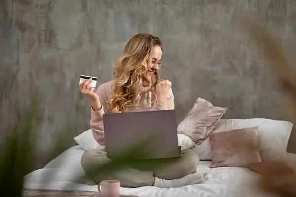 Loira modelo blogger em óculos, roupas casuais. Sorrindo, olhando animado, segurando cartão de plástico branco, sentado na cama com laptop nas pernas. Close-up — Fotografia de Stock